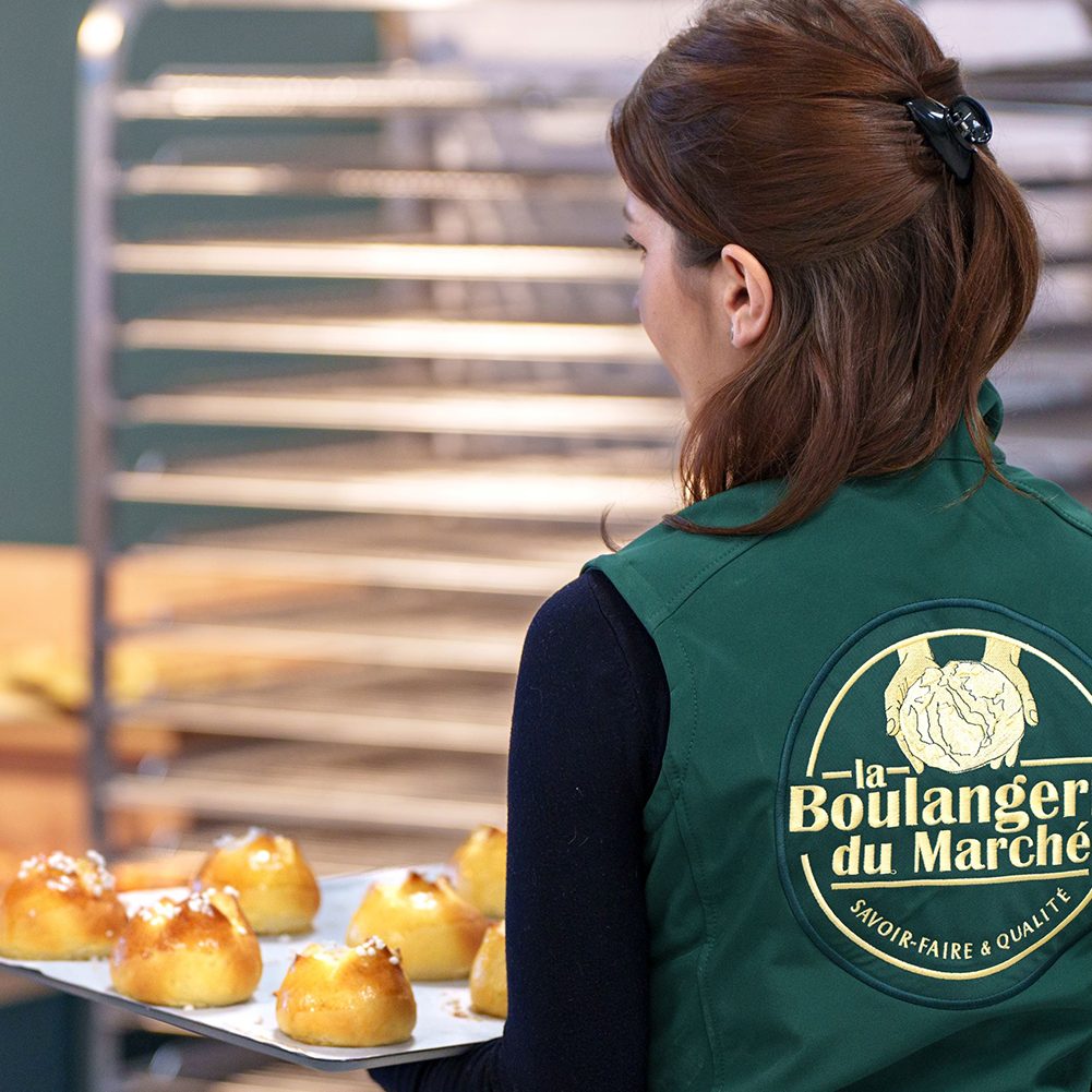 Un employé de la Boulangerie du Marché, vêtu d'un uniforme vert avec le logo "Savoir-faire & Qualité", portant un plateau de petits pains dorés fraîchement cuits, dans un environnement de boulangerie artisanale.